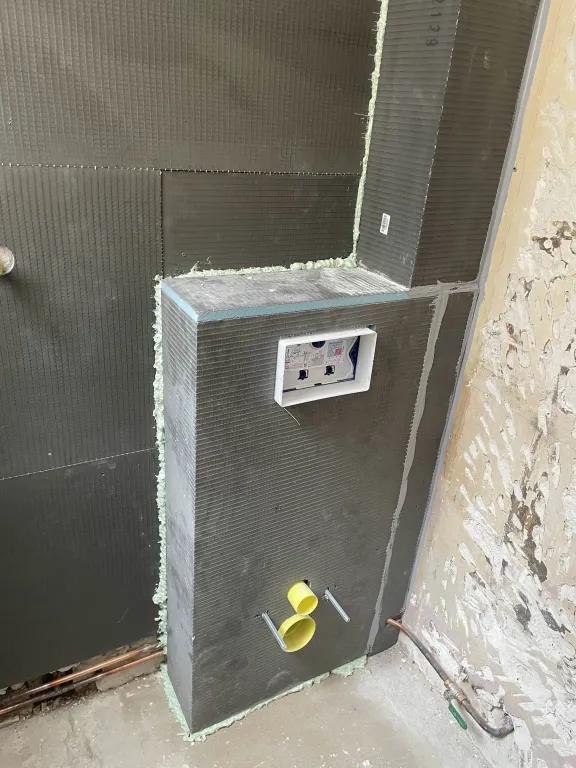 Vue rapprochée d'une installation de bâti-support pour WC suspendu dans une salle de bain en construction. Le bâti est entièrement coffré de panneaux d'isolation gris prêts à carreler (type Wedi), avec une ouverture rectangulaire pour la plaque de commande en haut et deux tuyaux jaunes raccordés en bas. Le mur de droite est brut et dégradé, tandis que le mur de gauche est également carrelé de panneaux. De la mousse d'étanchéité verte est visible sur la gauche.