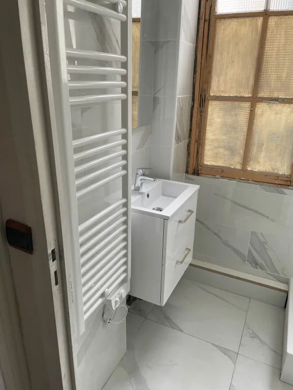 Salle de bain en cours de finition : un sèche-serviettes blanc au premier plan, un meuble-vasque suspendu blanc avec miroir au milieu, et des carreaux de marbre gris et blancs sur les murs. Une partie du mur reste non carrelée près d'une fenêtre en bois.