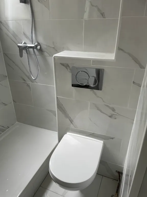 Intérieur de salle de bain moderne avec carrelage effet marbre blanc. À droite, un WC suspendu blanc avec une plaque de commande double chromée et une petite niche de rangement carrelée juste au-dessus. À gauche, un coin douche à l'italienne avec un receveur de douche blanc encastré et une colonne de douche en chrome avec flexible.